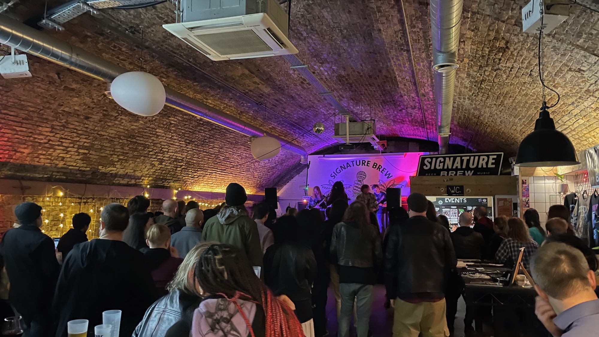 Photo of the interior of the Signature Brew Haggerston pub, showing a band on stage and a slew of people watching.